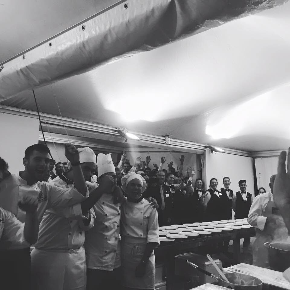 Chef e allievi esultano insieme in cucina, con i piatti pronti per il servizio sullo sfondo